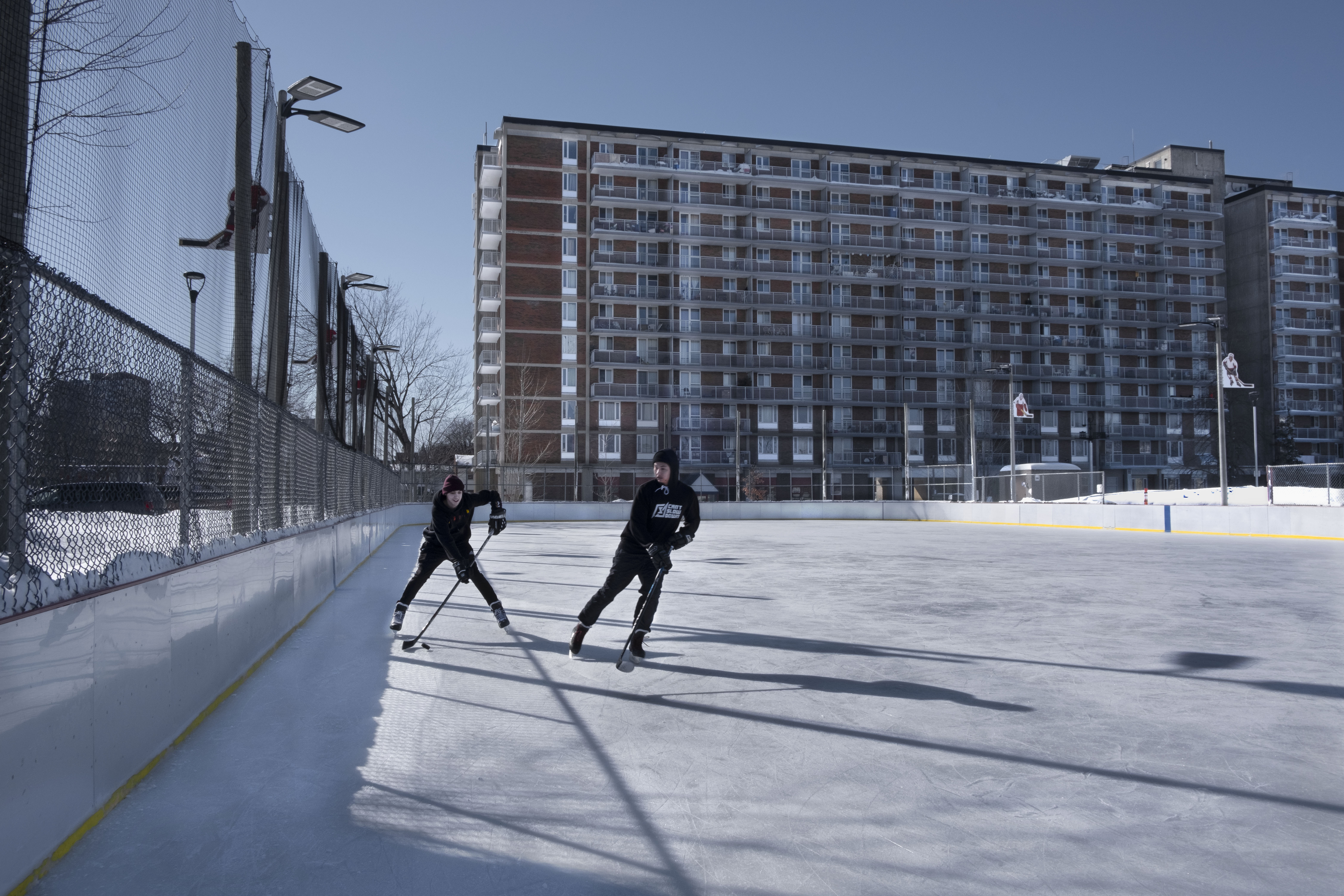 Two men play hockey on a community rink with a high rise apartment in the background.